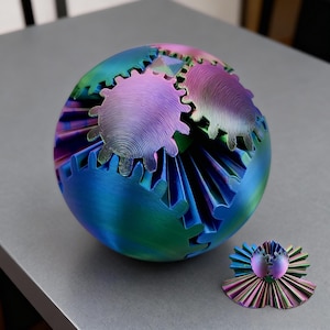 May include: An iridescent metal sphere sculpture with gear-shaped cutouts, predominantly blue, green, and purple. A smaller, matching piece sits beside it. The gear details give it a distinctive, industrial appearance. The sculpture is a decorative item.
