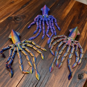 Può includere: Tre giocattoli di calamari colorati e articolati sono esposti su una superficie di legno. I calamari sono principalmente blu, con sfumature arcobaleno di giallo, arancione e verde. Ogni calamaro ha grandi occhi rotondi e molteplici tentacoli articolati.