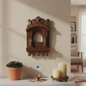 May include: A wall-mounted brown and gold decorative arch frame with an open centre, positioned above a surface featuring a small brass oil lamp, a terracotta potted succulent, and two cream-coloured pillar candles on a wooden stand.