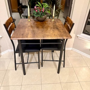 May include: A small dining set with a square, wood-grain table and two chairs. The table has a black metal frame and is topped with a decorative plant and candles. The chairs have black seats and wooden backs.