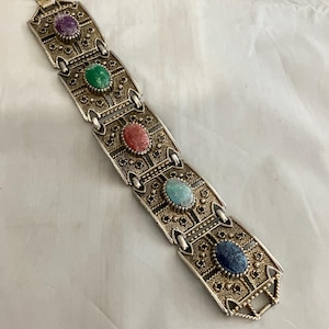 May include: Vintage silver-tone bracelet with ornate filigree design. The bracelet features five oval gemstones in purple, green, red, turquoise, and blue. The bracelet is approximately 7.5 inches long.
