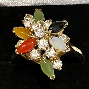 May include: A gold ring with a cluster of colorful gemstones, including green jade, orange, red, black, and white opal. The ring is set with clear rhinestones.