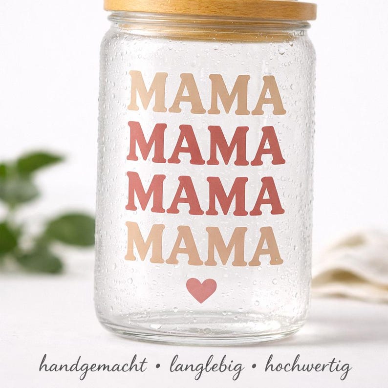 May include: A clear glass tumbler with a bamboo lid featuring the word MAMA printed four times in alternating tan and terracotta colors, stacked vertically, with a small pink heart icon below the text and water droplets on the glass surface.