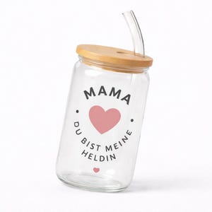 May include: A clear glass tumbler with a bamboo lid and a curved glass straw. The front of the glass features the black text MAMA, a pink heart, and the German text DU BIST MEINE HELDIN, followed by a small pink heart at the bottom of the design.