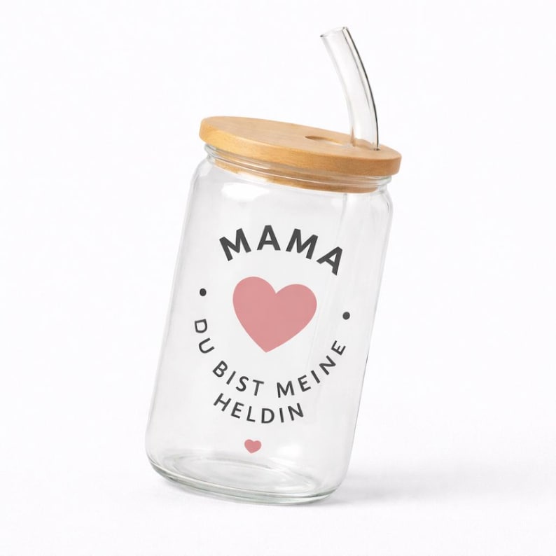 May include: A clear glass tumbler with a bamboo lid and a curved glass straw. The front of the glass features the black text MAMA, a pink heart, and the German text DU BIST MEINE HELDIN, followed by a small pink heart at the bottom of the design.