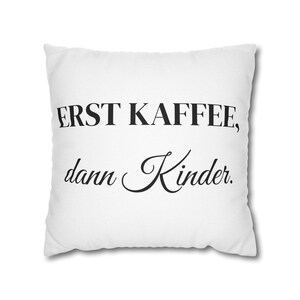 Może przedstawiać: Biała kwadratowa poduszka z napisem "ERST KAFFEE, dann Kinder" czarnym skryptem. Poduszka jest przedmiotem dekoracyjnym, prawdopodobnie na sofę lub łóżko, i ma prosty, nowoczesny design.