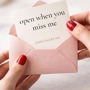 Op de afbeelding: Een lichtroze envelop bevat een kaart met de woorden "open when you miss me" in het zwart. De kaart zegt ook "A letter just for you". De envelop wordt vastgehouden door handen met rode nagellak.