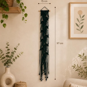 Op de afbeelding: Een donkergroene macramé wandhanger, 97 cm lang en 15 cm breed, hangt aan een beige muur. De handgemaakte textielkunst heeft een geweven ontwerp met geknoopte details en franjes. De decoratie is in boho-stijl.