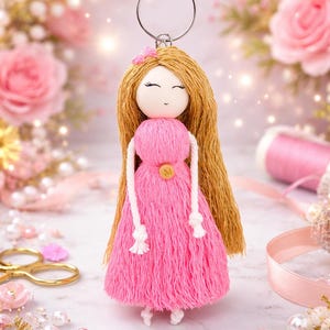 May include: A handmade doll keychain with long, golden hair and a pink dress. The doll has a sweet face and is adorned with a small pink bow. The keychain is made of yarn and has a metal ring for attaching to keys or a bag.