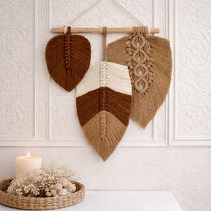 Könnte beinhalten: Makramee-Wandbehang mit drei blattförmigen Designs in Braun- und Beigetönen, aufgehängt an einer Holzdübel. Die Blätter weisen aufwendige Webmuster auf und verleihen jedem Raum einen Hauch von Boho-Dekor.