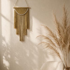 Può includere: Un arazzo da parete in macramè fatto a mano nei toni del beige e del marrone. Il design presenta una parte superiore a forma di semicerchio con un bordo di perline e frange a cascata. La decorazione murale è sospesa a un tassello di legno e un cordoncino in fibra naturale.