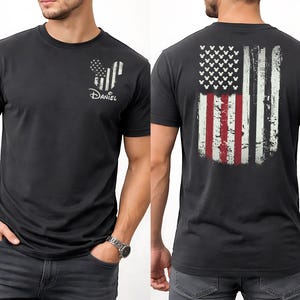K&ouml;nnte beinhalten: Dunkelgraues T-Shirt mit einem abgenutzten amerikanischen Flaggen-Design auf dem R&uuml;cken. Die Vorderseite zeigt eine von Mickey Mouse inspirierte Flaggen-Grafik und den Namen "Daniel" in Wei&szlig;. Das Shirt hat kurze &Auml;rmel und einen Rundhalsausschnitt.