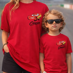 Pu&ograve; includere: Due magliette rosse con una grafica di un'auto dei cartoni animati. Una maglietta ha il nome "Olivia" e l'altra "Daniel" stampato sotto l'auto. Le magliette sono indossate da una donna e un bambino.