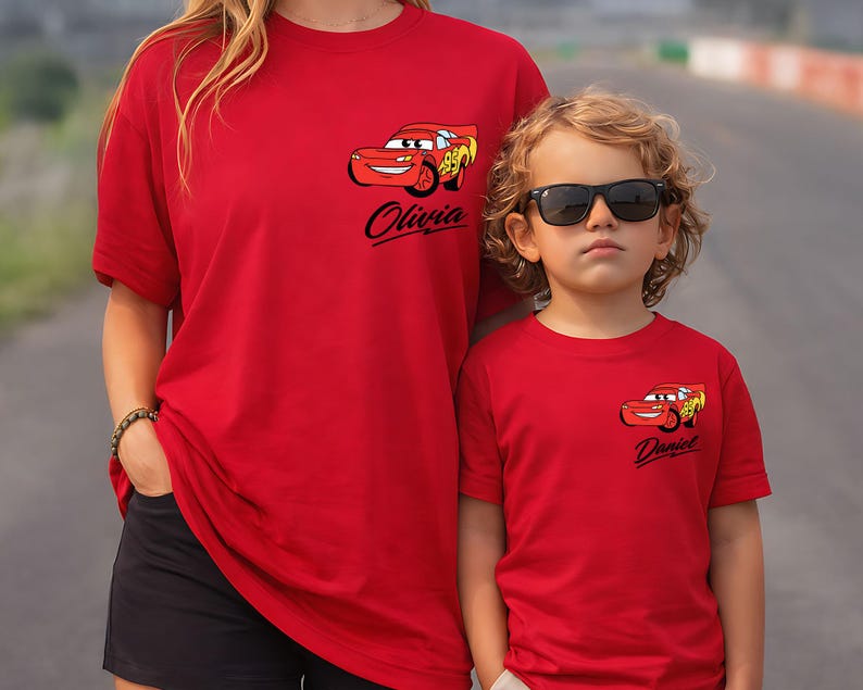 Pu&ograve; includere: Due magliette rosse con una grafica di un'auto dei cartoni animati. Una maglietta ha il nome "Olivia" e l'altra "Daniel" stampato sotto l'auto. Le magliette sono indossate da una donna e un bambino.