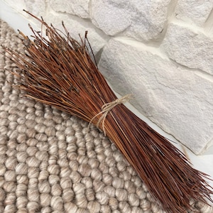 Puede incluir: Un manojo de agujas de pino secas, de color marrón rojizo, atadas con hilo. Las agujas están dispuestas en un grupo compacto, creando una estética rústica y natural. El manojo descansa sobre una alfombra beige texturizada, con una pared de piedra blanca al fondo.