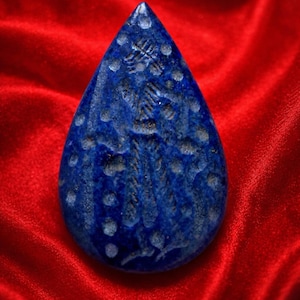 May include: A teardrop-shaped, deep blue lapis lazuli pendant with white speckles and carved details. The stone is set against a vibrant red background, highlighting its colour and texture. The pendant's surface features intricate carvings.