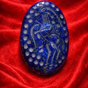 May include: Oval-shaped, deep blue lapis lazuli pendant with a carved design of a figure with a bow and arrow. The pendant has a border of white dots and is set against a red velvet background.