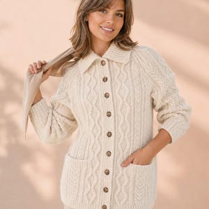 Könnte beinhalten: Eine cremefarbene Strickjacke mit Zopfmuster, Kragen, langen Ärmeln und einer Knopfleiste mit braunen Knöpfen. Das Kleidungsstück verfügt über zwei aufgesetzte Vordertaschen und ein durchgehendes, strukturiertes vertikales Zopfmuster.