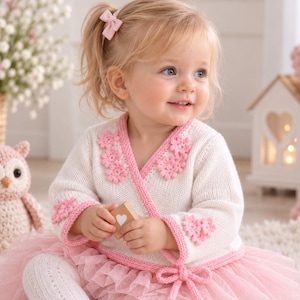 Peut inclure: Un gilet cache-cœur en tricot crème orné d'applications florales et en forme de cœur roses, associé à une jupe en tulle rose superposée et des collants blancs à motifs. Le gilet présente un liseré rose et un lien latéral, avec des chaussures roses.