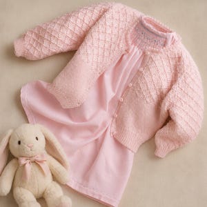 May include: A light pink knitted cardigan with a diamond texture pattern and button closure, paired with a matching light pink dress featuring a smocked neckline. A cream-colored plush rabbit toy with a pink ribbon bow sits beside the clothing.