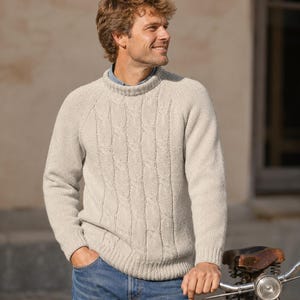 May include: A cream-colored cable knit sweater with a ribbed crew neckline, long sleeves, and ribbed cuffs. The sweater features a vertical cable pattern on the front panel and is worn over a light blue collared shirt with blue denim jeans.