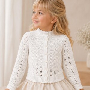 May include: A white long-sleeved knit cardigan featuring a textured pattern, a high neckline, and a vertical row of small matching buttons down the front. The garment has ribbed cuffs and a ribbed hem, providing a structured fit for the upper body.