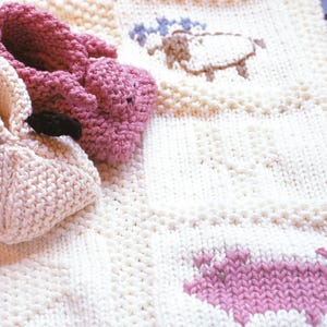 Puede incluir: Primer plano de una manta y patucos de bebé tejidos. La manta es de color blanco roto con un diseño de oveja en marrón y azul. Los patucos son rosas y blanco roto, con un detalle negro. Los artículos están hechos de un hilo suave y texturizado.