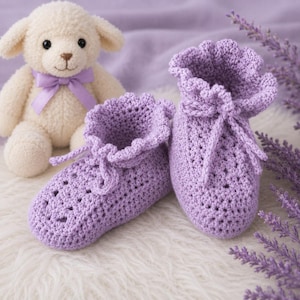 May include: A pair of light purple crocheted baby booties featuring a ruffled top edge and a matching tie closure. The booties are crafted with a textured stitch pattern and are positioned next to a cream-colored plush lamb toy.