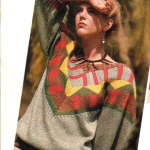 May include: A gray sweater with a geometric pattern in red, yellow, and green. The sweater has long sleeves and a crew neck. The design is on the chest and sleeves. The model is wearing a necklace.