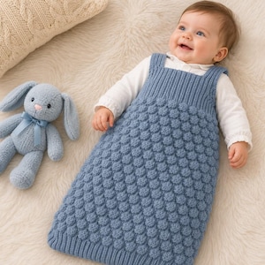 Peut inclure: Un bébé dans un sac de couchage bleu tricoté avec un motif de bulles texturées. Le bébé porte une chemise blanche à manches longues. Un lapin en peluche bleu avec un ruban bleu est à côté du bébé. Le bébé est allongé sur une surface blanche et moelleuse.