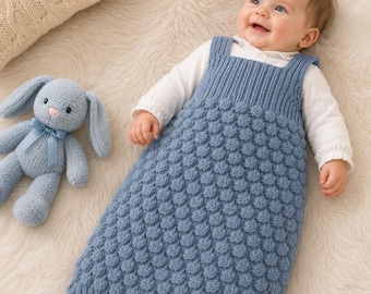 May include: A baby wearing a white long-sleeved shirt under a dusty blue knitted sleeveless sleep sack featuring a textured bobble stitch pattern and ribbed trim, accompanied by a matching dusty blue plush rabbit toy with a blue ribbon.