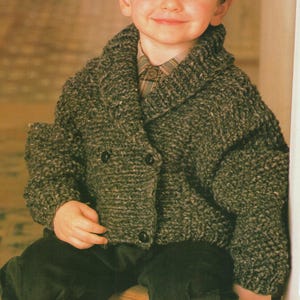 May include: A child wearing a chunky, gray knit jacket with a shawl collar and two dark buttons. The jacket is paired with a plaid shirt and dark corduroy pants. The jacket appears to be handmade.