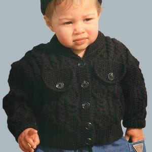 May include: A black knitted baby cardigan with a cable knit design. The cardigan has a button-up front and two pockets. The baby is also wearing a black hat and blue jeans.