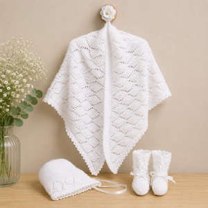 Può includere: Un set neonato bianco lavorato a maglia, composto da uno scialle, un cappello e delle scarpine. Lo scialle presenta un motivo a rombi ed è appeso a un piolo di legno con un fiore. Il cappello ha un bordo smerlato e nastri.