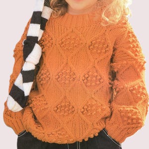 Puede incluir: Un suéter tejido de color naranja con un patrón de diamantes y bolitas texturizadas. El suéter de manga larga tiene cuello redondo y se combina con una bufanda a rayas blancas y negras. El suéter está hecho de un material suave y cálido.