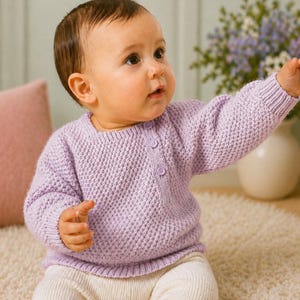以下が含まれることがあります： 淡い紫色のベビーセーター。 質感のあるニット模様です。 セーターはクルーネック、長袖、3つのボタン留めです。 赤ちゃんは白いズボンを履いています。