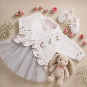 May include: A white knitted long-sleeve dress with a tulle skirt, featuring a scalloped collar and cuffs embroidered with small pink rosebuds. The set includes a pair of white knitted booties with ribbon ties and a plush beige rabbit toy.