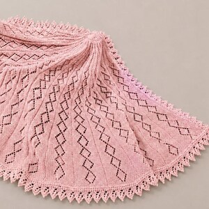 May include: A light pink knitted shawl with a diamond pattern and a scalloped edge. The shawl is made of a soft, lightweight material and is draped over a surface. The design features openwork diamonds and a delicate border.