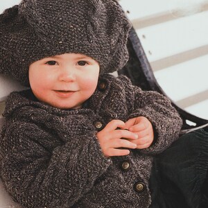 May include: A child wearing a dark gray knitted beret and matching button-up jacket. The jacket has decorative buttons. The knitted outfit appears to be warm and cozy, suitable for cooler weather. The child is smiling.