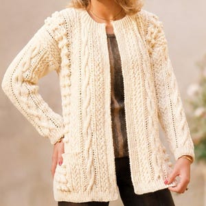 Peut inclure: Un cardigan ouvert de couleur blanc cassé à manches longues, orné d'un motif complexe en tricot torsadé, de détails côtelés verticaux et de pompons texturés. Le vêtement comprend deux poches plaquées à l'avant et une encolure ronde côtelée.