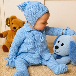 May include: A baby wearing a light blue knitted outfit, including a hat with a tassel, a button-up jacket, and matching pants. The baby is interacting with a blue stuffed dog. A brown teddy bear is in the background.
