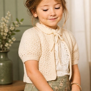 May include: A cream-colored, short-sleeved knitted cardigan with a round collar and a tie closure. The cardigan is worn over a white blouse with a lace trim and a floral patterned skirt. A gold bracelet is visible on the wrist.