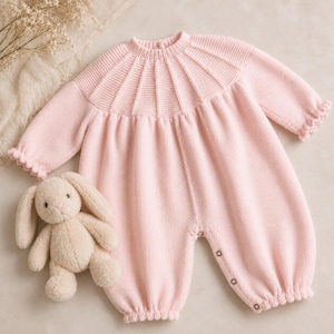 May include: A light pink knitted romper featuring a ribbed round neckline with a textured yoke pattern, long sleeves with ruffled cuffs, and a buttoned leg opening, accompanied by a small, beige plush rabbit toy with long ears.