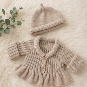 May include: A beige knitted baby outfit, comprising a cardigan with wooden buttons and a matching beanie hat. The cardigan features a ruffled hem and a rounded collar. The set is presented on a soft, white, textured surface.