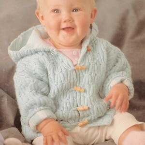 May include: A light blue knitted baby hooded cardigan with wooden toggle closures. The sweater has a textured pattern and ribbed cuffs. The baby is wearing a pink shirt and white pants. The baby is smiling.