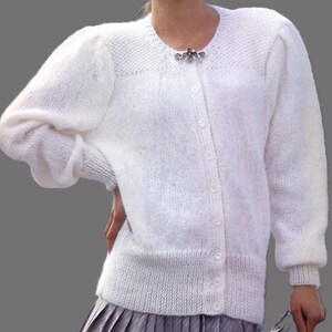 May include: A white cardigan with a round neck and long sleeves. The knitted sweater has a textured pattern and a decorative brooch near the neckline. The sleeves have a slight puff at the shoulders. The cardigan is buttoned down the front.