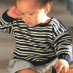 May include: A navy blue and white striped sweater with a round neckline. The sweater has long sleeves and is made of a knitted fabric. The child is also wearing striped shorts.