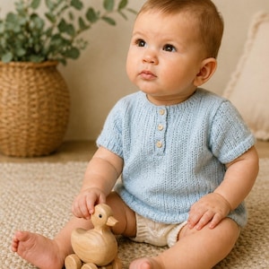 May include: A baby in a light blue knitted top with three wooden buttons, paired with white shorts, holding a wooden duck toy. The baby is seated on a beige rug. The top is a soft, knitted material.
