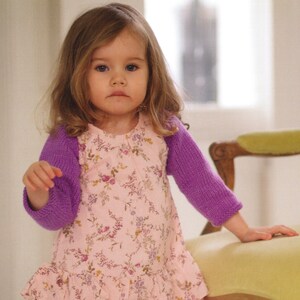 May include: A young child wearing a pink floral dress with a purple knitted shrug. The dress has a ruffled hem and a delicate floral pattern. The shrug covers the arms and shoulders.