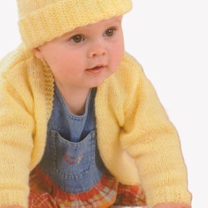 Puede incluir: Un bebé con gorro y cárdigan de punto amarillo sobre un peto vaquero con falda a cuadros. El bebé mira hacia un lado. La ropa de punto es de color amarillo suave.